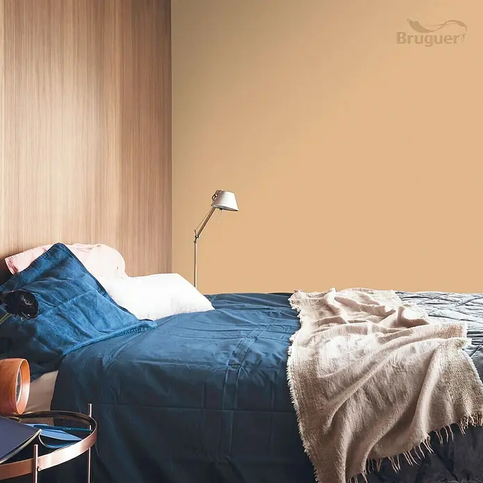 Bruguer Colores del Mundo Tester de pintura - Egipto ocre natural, Mate, 30 ml - Pared de dormitorio pintada con pintura Bruguer, creando un ambiente cálido y acogedor. Cama con ropa de cama azul y cojines claros.