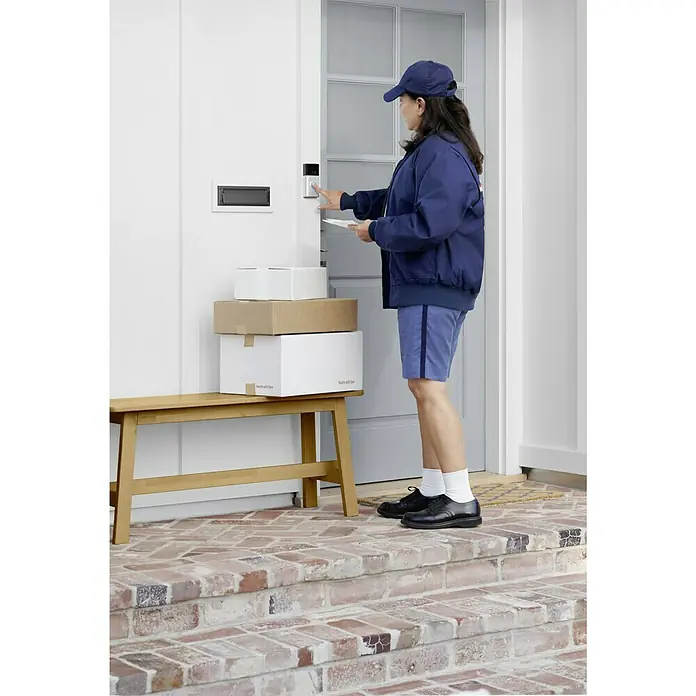 Ring Timbre con cámara Video Doorbell - Niquel - Una mujer en uniforme de reparto está usando un timbre Ring junto a paquetes apilados en un banco frente a una puerta principal.