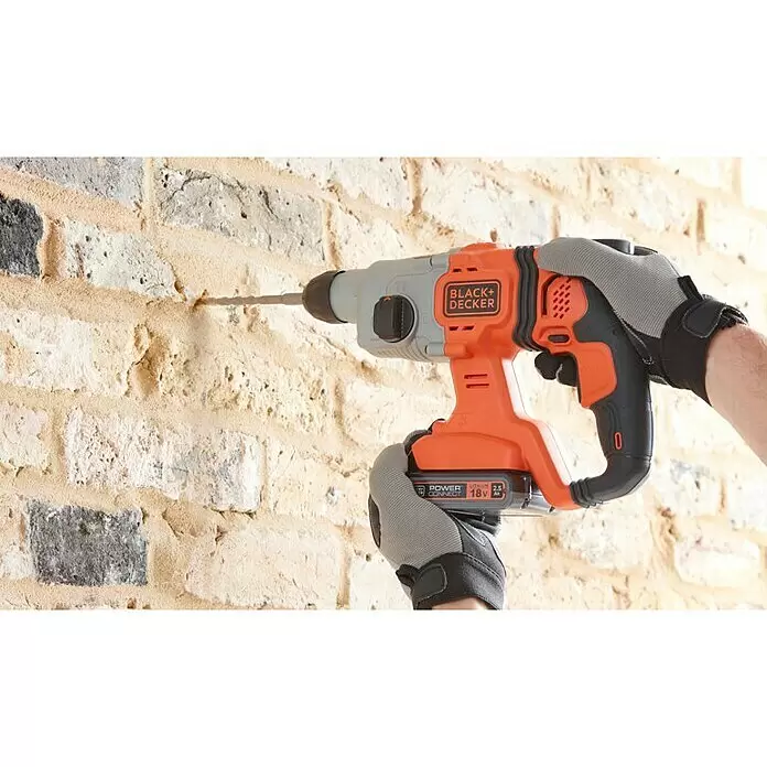 Black+Decker Martillo combinado de batería BCD900E2K - 18 V, 2 baterías, 1,2 J - Taladro percutor Black+Decker naranja y negro, sujetado por manos enguantadas, perforando activamente una pared de ladrillo claro.