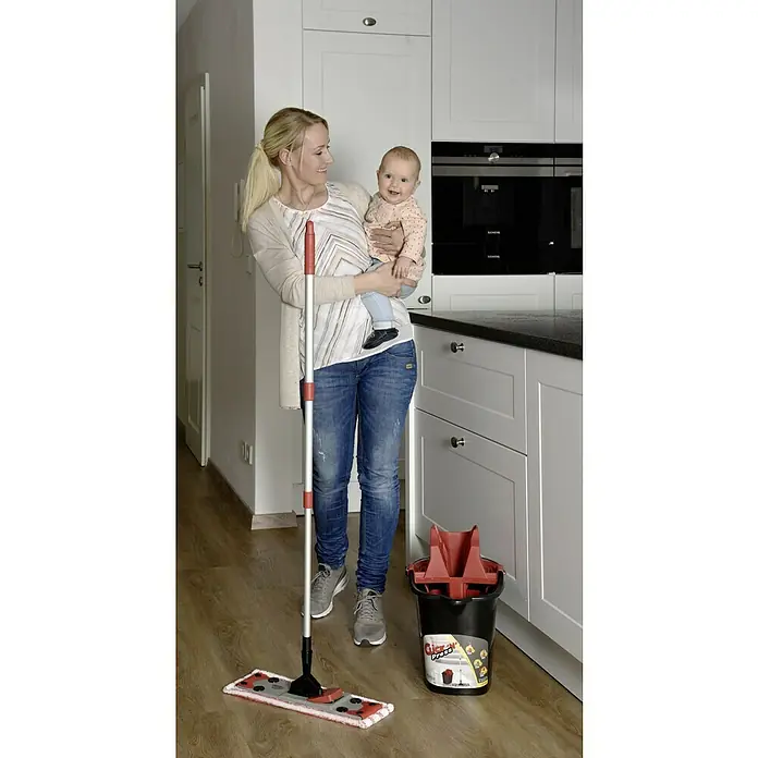 Sprintus Bodenwisch-Set Click N Press + Staubmop - 12 l, 5 -tlg., Materialzusammensetzung Bezug: 80 % Mikrofaser, 20 % weiche Abrasivborsten - Eine Frau hält ein Baby und steht neben einem Wischmopp und einem Eimer Click 'N' Press.