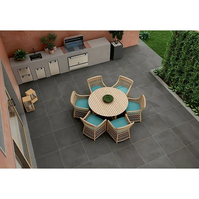 BHS Showroom Pavimento porcelánico Rodano - 60 x 60 cm, Antracita, Rectificado - Una zona al aire libre con una mesa redonda de madera y sillas de mimbre con cojines azules, rodeada de baldosas oscuras.
