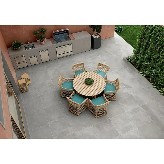 Pavimento porcelánico Rodano - 75 x 75 cm, Gris oscuro, Rectificado, C-3 Antideslizante - Esta imagen muestra un elegante diseño de patio con una mesa redonda de madera y sillas de mimbre con cojines turquesas.