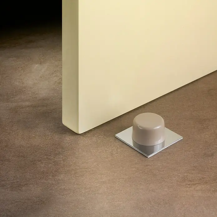 Inofix Tope de puerta 2023 6 uds. - Plástico, Beige - Un tope de puerta de metal y plástico sujeto a una puerta amarilla clara y un suelo marrón, mostrando su diseño.