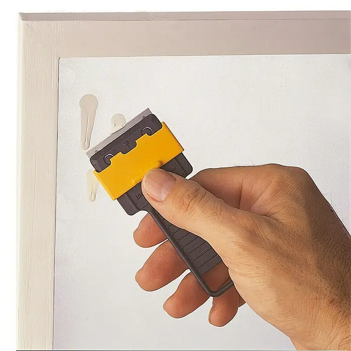 Inofix Juego de cuchillas de repuesto - 5 ud. - Se está utilizando un raspador manual para quitar pintura seca de una superficie blanca. Perfecto para limpiar superficies delicadas suavemente.