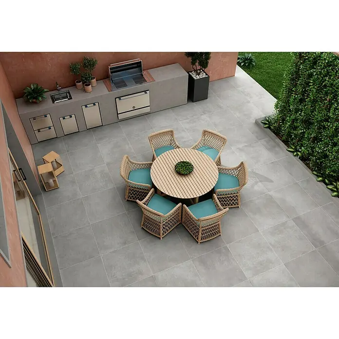 BHS Showroom Pavimento porcelánico Rodano - 60 x 60 cm, Gris oscuro, Rectificado - Una mesa redonda de madera con seis sillas de mimbre y cojines turquesa en un patio exterior gris.