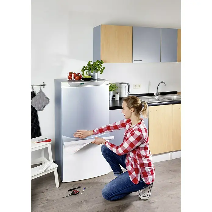 D-c-fix Metalleffektfolie Metallic - 150 x 67,5 cm, Silber, Selbstklebend - Frau bringt eine selbstklebende Folie an einem Kühlschrank an, um die Küche individuell zu gestalten. Küchenrenovierung leicht gemacht!