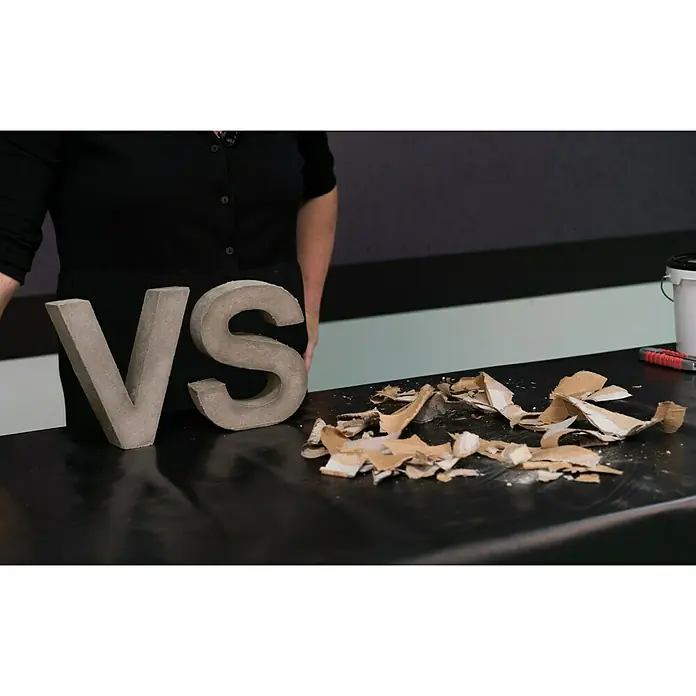 Ultrament Beton-Gießmasse Bastel Beton - Hellgrau, 1,5 kg - Bild zeigt die Betonbuchstaben VS mit der Person, die diese hält und mit Pappe auf dem Tisch vor dem Hintergrund mit grauen und schwarzen Streifen.