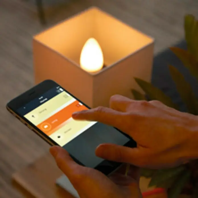 Philips Hue Smart-LED-Lampe White Ambiance - E14, Dimmbar, Warmweiß, 470 lm, 5,2 W - Eine Nahaufnahme eines Smartphones, das eine App zur Steuerung einer intelligenten Glühbirne anzeigt, die in einem würfelförmigen Lampenschirm sitzt.