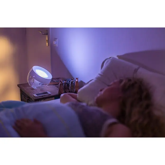 Philips Hue LED-Tischleuchte Iris - Weiß/Transparent, Höhe: 19,4 cm - Intelligente Lampe auf Nachttisch mit schlafender Person im Bett. Die smarte Lampe erhellt den Raum mit einem beruhigenden Schein.