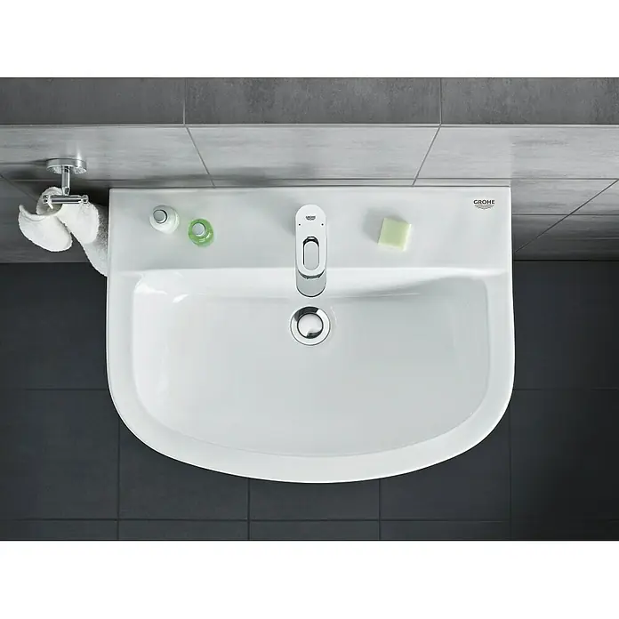 Grohe BauLoop Waschtischarmatur S-Size - Chrom, Glänzend - Aufsicht auf ein GROHE Waschbecken im Badezimmer mit Armatur, Seife und Handtüchern. Das Waschbecken ist weiß und die Fliesen sind grau.