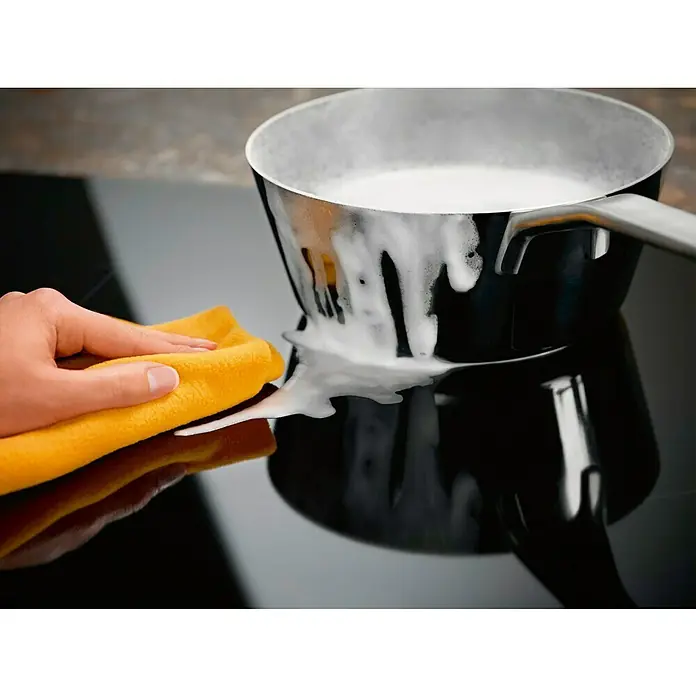Electrolux Placa de cocción LIT60336C - Número de fogones de inducción: 3 ud. - Un paño amarillo limpia leche sobre una placa de inducción negra alrededor de una olla.