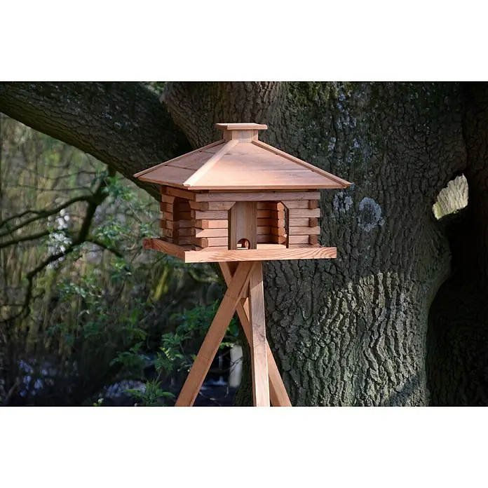 Dobar Vogelhaus Rustikal - 50 x 50 x 38 cm, Holz, Hellbraun - Nahaufnahme eines hölzernen Vogelhauses auf einem Ständer, mit Baum im Hintergrund. Das Vogelhaus hat ein einzigartiges Design mit mehreren Eingängen.