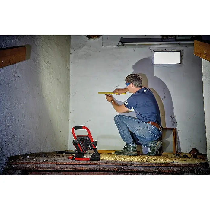 Einhell PXC 18V Foco de batería LED TC-CL 18 - 18 V, Sin batería, Flujo luminoso: 1.800 lm - Un artesano mide una pared con un aparato de medición amarillo, iluminado por una luz de trabajo roja.