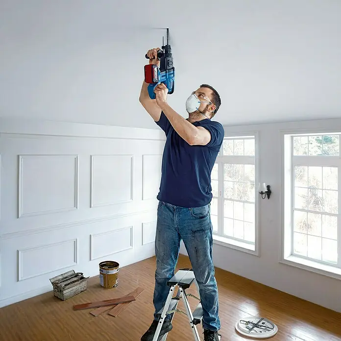 Bosch Professional AMPShare 18V Martillo combinado de batería GBH 18V-21 - 18 V, 2 baterías, 4 Ah, 2 J, Maletín incluido - Un hombre usa un taladro percutor inalámbrico Bosch para taladrar el techo mientras está parado en una escalera y usa una máscara.