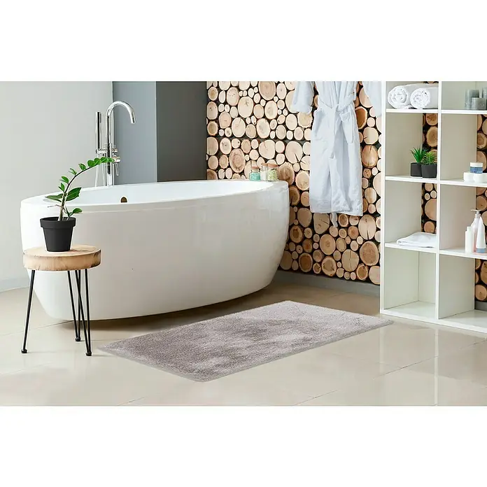 Badteppich Happy - 50 x 90 cm, Silber, 100% Polyester - Ein grauer, flauschiger Badezimmerteppich liegt vor einer modernen Badewanne und einem Holzscheiben-Akzentwand. Ideal für ein stilvolles Badezimmer.
