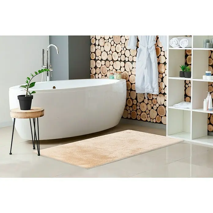Badteppich Happy - 67 x 110 cm, Beige, 100% Polyester - Eine luxuriöse, beige Badematte liegt auf dem hellen Fliesenboden vor einer freistehenden Badewanne in einem stilvollen Badezimmer.