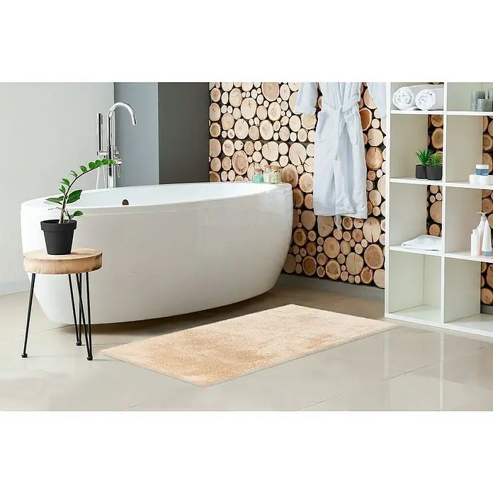 Badteppich Happy - 50 x 90 cm, Beige, 100% Polyester - Ein beigefarbener Badteppich liegt vor einer Badewanne mit Holzscheibenwand, neben einem Regal und einem kleinen Tisch mit Pflanze.