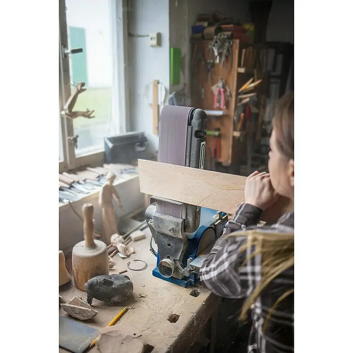 Scheppach Band- & Tellerschleifer BTS900 - 370 W, 2.850 U/min - Nahaufnahme eines Bandschleifers mit einer Person, die ein Holzstück schleift, in einer Werkstatt mit Holzschnitzereien und Werkzeugen.