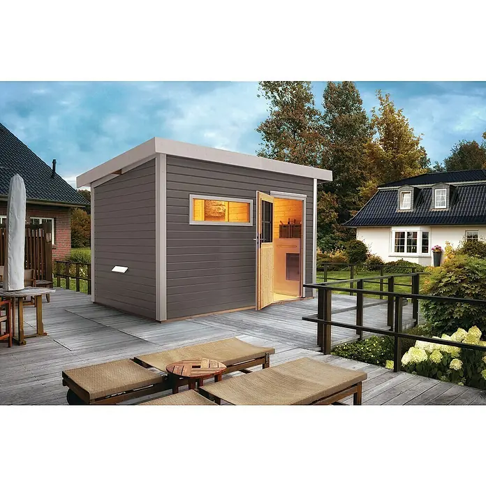Karibu Saunahaus Suva 2 - Mit 9 kW Bio-Ofen mit externer Steuerung, Terragrau, L x B x H: 231 x 337 x 227 cm - Ein modernes Gartenhaus mit grauem Holz und offenstehender Tür, das auf einer Holzterrasse mit Liegestühlen steht, ideal für den Außenbereich.