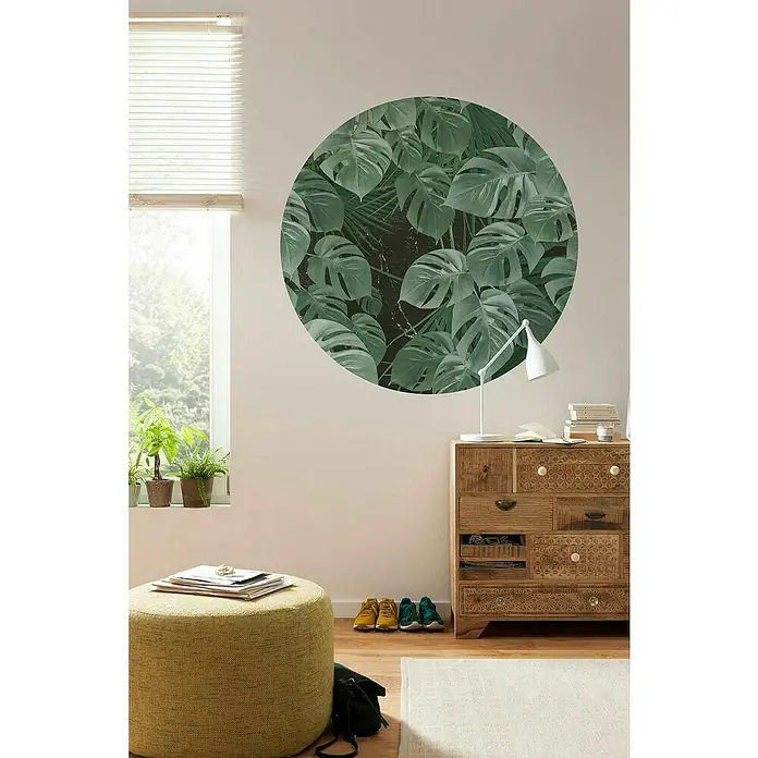 Komar Dots Fototapete rund Novel Monstera - 125 cm, Selbstklebend - Ein Interieur mit einem runden Wandbild aus Monstera-Blättern über einer Holzkommode, die mit einer Lampe und Büchern dekoriert ist.