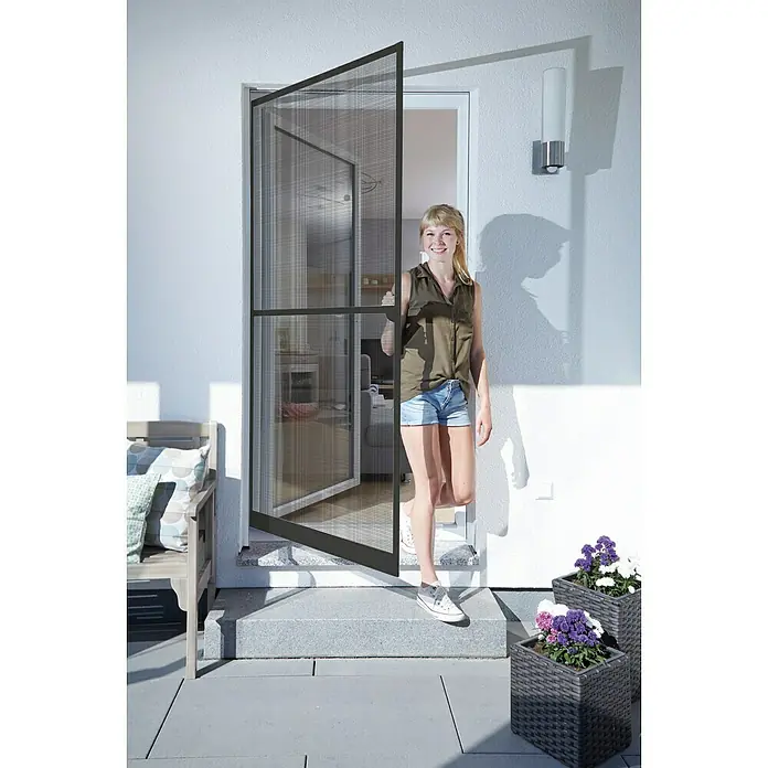 Schellenberg Mosquitera Action - An x Al: 100 x 210 cm, Montaje de la mosquitera: Atornillado, Color bastidor: Antracita - Una mujer está frente a una puerta mosquitera abierta. La puerta es negra y tiene una malla fina.