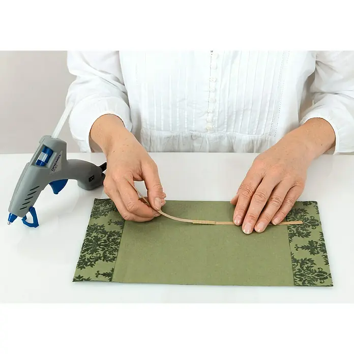 Dremel Barra de cola multiuso GG 11 - 11 mm, Específico para: Pistola termoadhesiva mod. 940, 12 ud. - Una persona usa una pistola de pegamento para fijar una tira artesanal en un sobre verde decorado que está sobre una mesa.