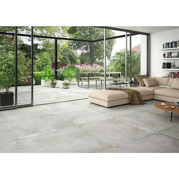 BHS Showroom Pavimento porcelánico Stoneage Soft - 100 x 100 cm, Concreto, Rectificado - Interior de una sala de estar con baldosas grises y vistas a un jardín con mesa y sillas de comedor.