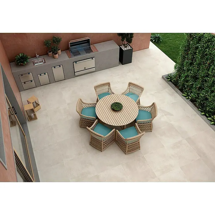 Pavimento porcelánico Rodano - 75 x 75 cm, Tiza, Rectificado - Una elegante zona de comedor al aire libre con una mesa redonda y sillas de mimbre con cojines turquesa, complementada con una cocina moderna y exuberante vegetación.