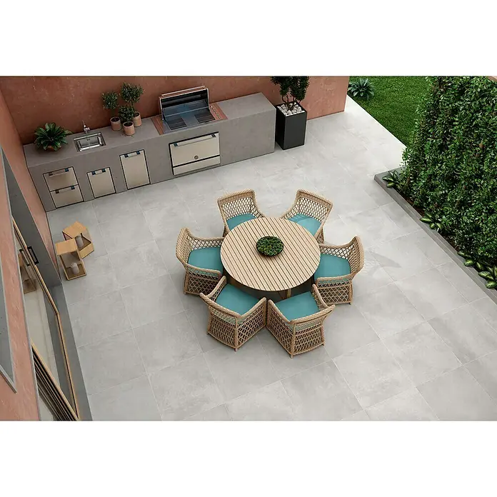BHS Showroom Pavimento porcelánico Rodano - 75 x 75 cm, Gris claro, Rectificado - Vista superior de una mesa de comedor al aire libre con sillas de mimbre y cojines turquesa, rodeada de una cocina exterior y vegetación.