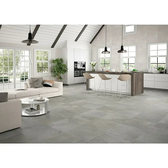 Pavimento porcelánico Rodano - 75 x 75 cm, Gris oscuro, Rectificado - Una sala de estar moderna y luminosa con suelos de baldosas grises, un sofá blanco, una mesa de centro y una isla de cocina con taburetes.