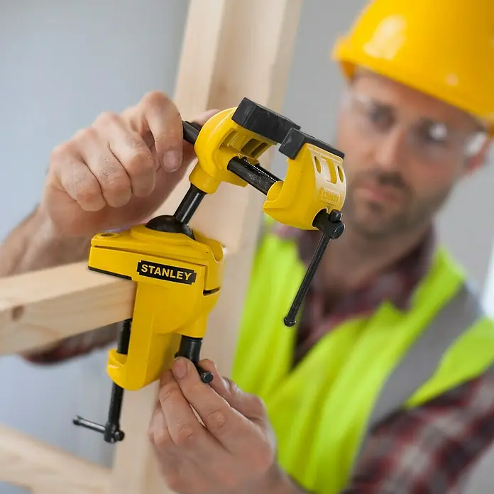 Stanley Tornillo de banco - Anchura de mordaza: 70 mm - El tornillo de banco amarillo Stanley está fijado a una pieza de madera por un trabajador con casco y gafas de seguridad.