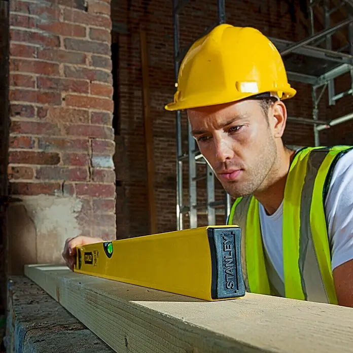 Stanley Nivel de burbuja STHT1-43102 - Aluminio, Amarillo - Un constructor usa un nivel Stanley amarillo para comprobar la planitud de un tablón de madera en una obra.