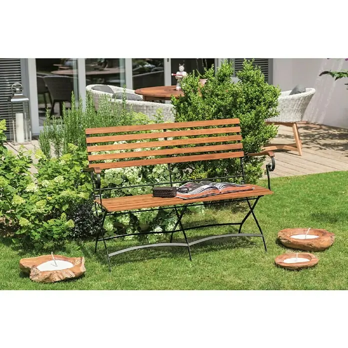 Sunfun Moni Banco de jardín - An x Pr x Al: 120 x 57 x 90 cm, Madera de acacia, Marrón natural/Negro, Abatible - Un banco de jardín de madera con estructura de metal negro está situado sobre un césped verde. Hay un libro abierto y una caja pequeña encima.