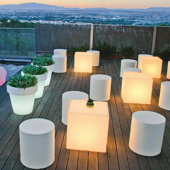 New Garden Design-Außenleuchte Cuby 32 - 32 x 32 x 32 cm, Warmweiß - Eine stimmungsvolle Szene mit beleuchteten Outdoor-Möbeln, darunter Würfel und Zylinder, die eine Terrasse mit Blick auf die Stadt bei Dämmerung erhellen.