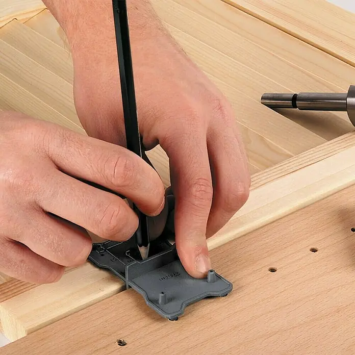 Wolfcraft Set de brocas Forstner - 2 pzs., Diámetro vástago: 10 mm - Una persona utiliza una plantilla de perforación y un lápiz para marcar agujeros precisos en una pieza de madera. La herramienta asegura un espaciado uniforme.