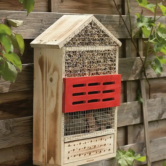 Windhager Insektenhotel - L x B x H: 33,5 x 14 x 49,5 cm, Holz, Naturbraun/Rot, Geflammt - Ein hölzernes Insektenhotel mit einem roten Akzent hängt an einem Holzzaun, umgeben von grüner Vegetation und bietet Unterschlupf für Insekten.