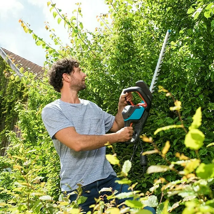 Gardena ComfortCut Cortasetos eléctrico 550/50 - 550 W, Longitud de corte: 50 cm, Grosor de rama máx.: 27 mm - Un hombre usa un cortasetos Gardena para cortar un seto. La herramienta es negra, turquesa y naranja para un corte preciso. Ideal para jardinería.
