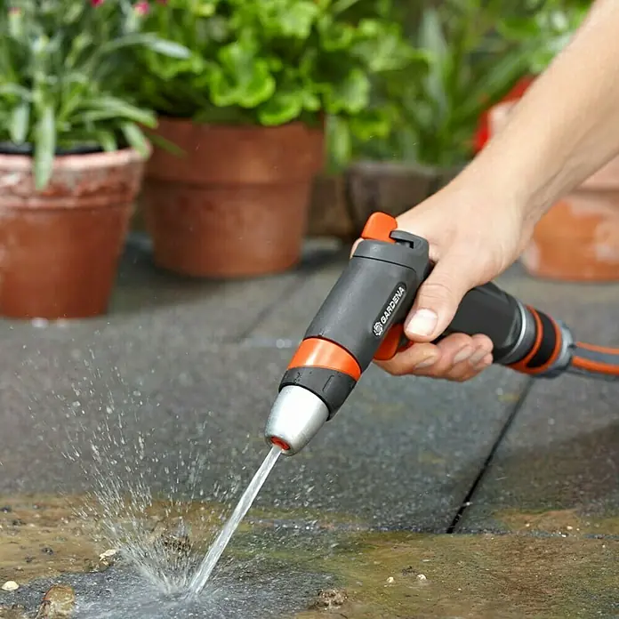 Gardena Premium Reinigungsspritze - Anzahl Sprühformen: 3 Stk., Metall/Kunststoff - Eine Hand hält eine Gardena-Spritzpistole, die einen Wasserstrahl auf eine Steinfläche richtet, mit Pflanzen im Hintergrund.