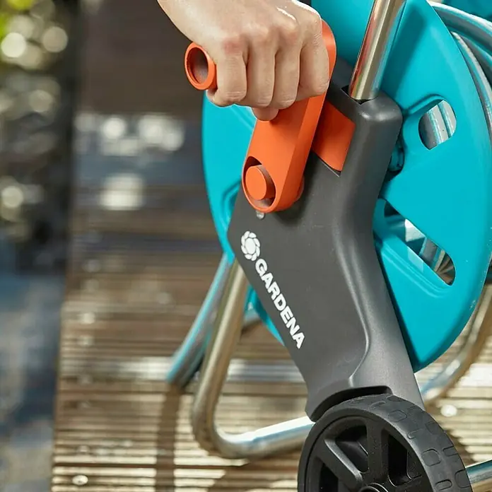 Gardena Schlauchwagen-Set CleverRoll S - Kunststoff/Metall, Schlauchlänge: 20 m - Nahaufnahme einer Hand, die eine Gardena Schlauchbox auf einem Holzdeck bedient. Der Schlauchwagen ist blau mit orangefarbenen Akzenten.