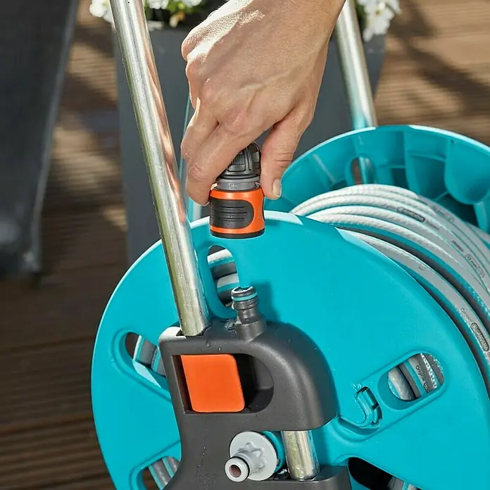 Gardena Schlauchwagen-Set CleverRoll S - Kunststoff/Metall, Schlauchlänge: 20 m - Nahaufnahme einer Person, die einen Schlauchanschluss an einer Gardena Gartenschlauchbox befestigt. Das blaue Gehäuse und der graue Schlauch sind gut sichtbar.