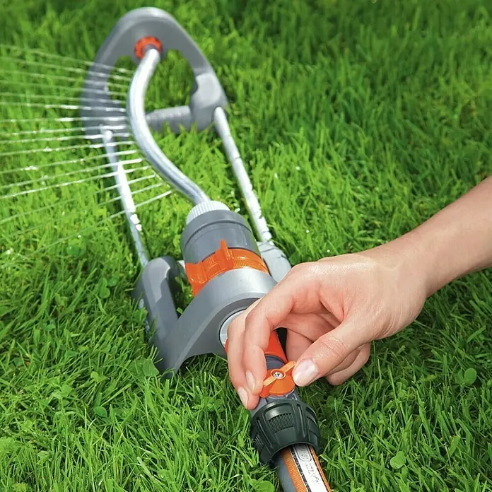 Gardena Regulierstop - ¾″ / ⁵⁄₈″ - Eine Person bedient einen Gardena Regner auf einem grünen Rasen, um den Garten zu bewässern. Der Regner ist grau mit orangefarbenen Akzenten.