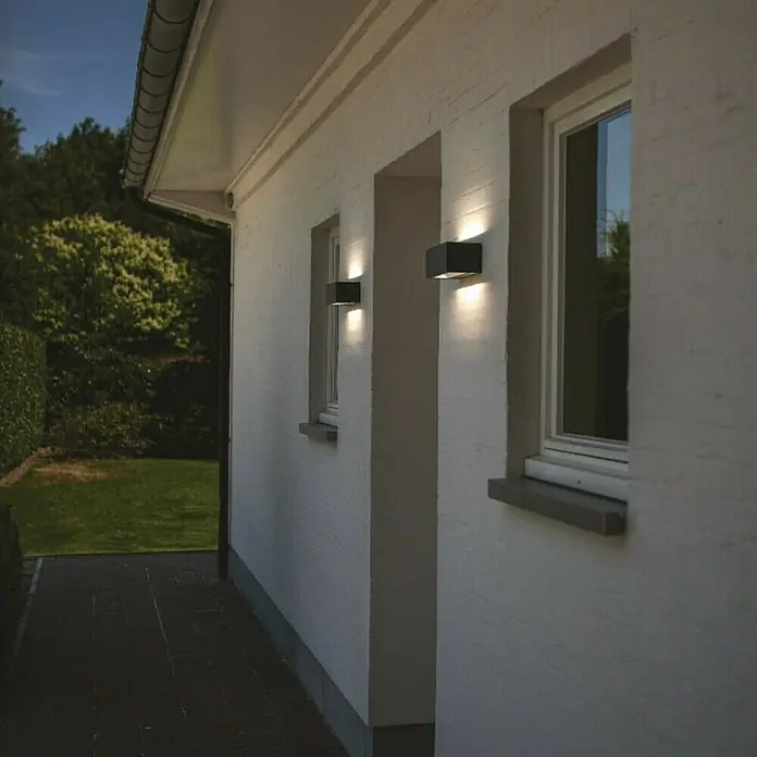 Lutec Aplique exterior LED Gemini - 20 W, 10,3 x 22 x 8,5 cm, Antracita, IP54 - Luces de pared exteriores modernas iluminan la pared blanca de una casa junto a ventanas y un camino pavimentado.