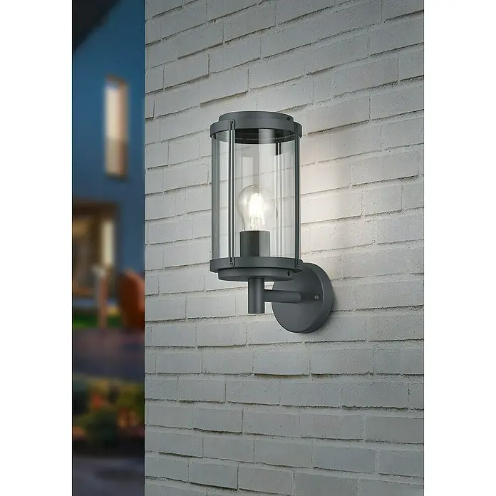 Trio Lighting Aplique exterior Tanaro - 40 W, L x An x Al: 18,5 x 14,5 x 32,5 cm, Antracita, IP44 - Una moderna lámpara de pared exterior con vidrio transparente y marco de metal gris ilumina una pared de ladrillos.