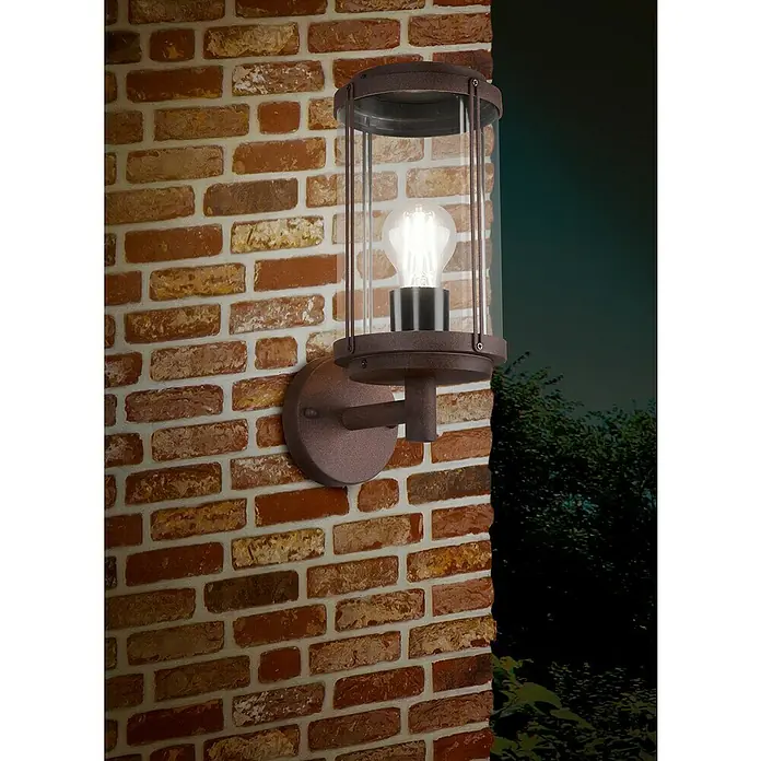 Aplique exterior Tanaro - 40 W, 14 x 20 x 31 cm, Marrón, IP44 - Un primer plano de un farol de pared exterior color óxido con cristal cilíndrico transparente y una bombilla Edison, montado en una pared de ladrillo.