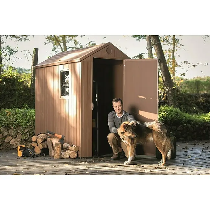 Keter Caseta de jardín Darwin 4x6 - Medidas ext. (An x Pr): 125,8 x 184,5 cm, Plástico, Marrón, 2,3 m² - Un hombre y su perro posan frente a un cobertizo de jardín espacioso y marrón, ideal para guardar cosas del jardín.
