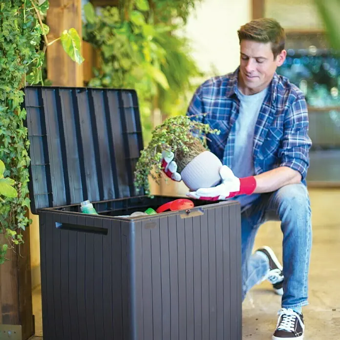 Keter Gartenbox City Box - L x B x H: 44 x 57,8 x 55 cm, Kunststoff, Anthrazit - Ein Mann, der neben einer grauen Aufbewahrungsbox kniet, während er eine Pflanze in einen Topf hält.