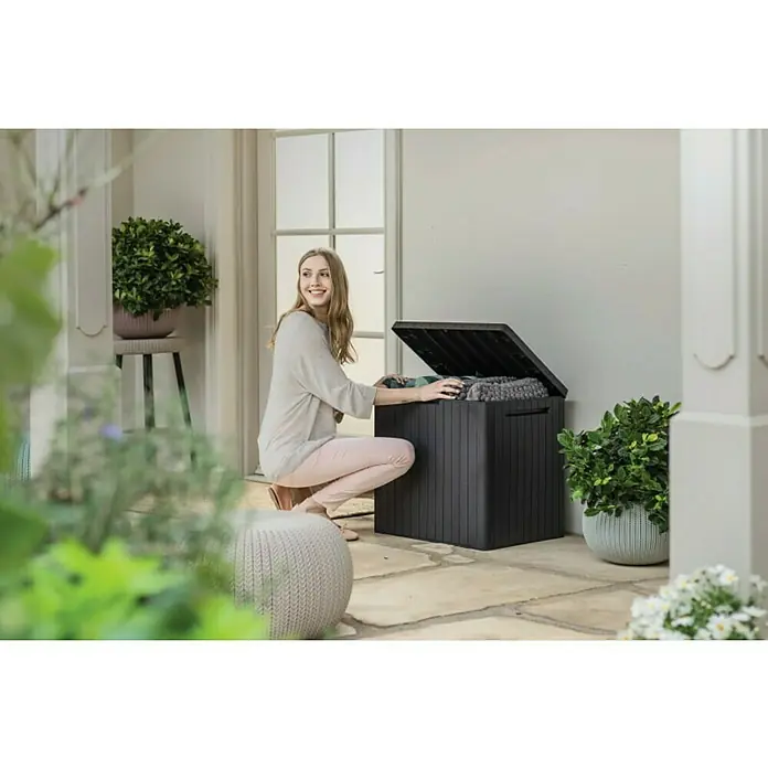 Keter Gartenbox City Box - L x B x H: 44 x 57,8 x 55 cm, Kunststoff, Anthrazit - Eine Frau kniet neben einer schwarzen Aufbewahrungsbox im Freien und verstaut Decken und Kissen.