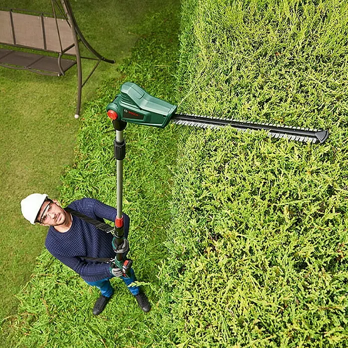 Bosch Power for All 18V Teleskop-Heckenschere UniversalHedgePole 18 - 18 V, Li-Ionen, Ohne Akku, Schnittlänge: 43 cm - Ein Mann, der eine Bosch Hochentaster-Heckenschere benutzt, um eine hohe Hecke zu schneiden, mit Sicherheitsausrüstung wie Helm und Brille.