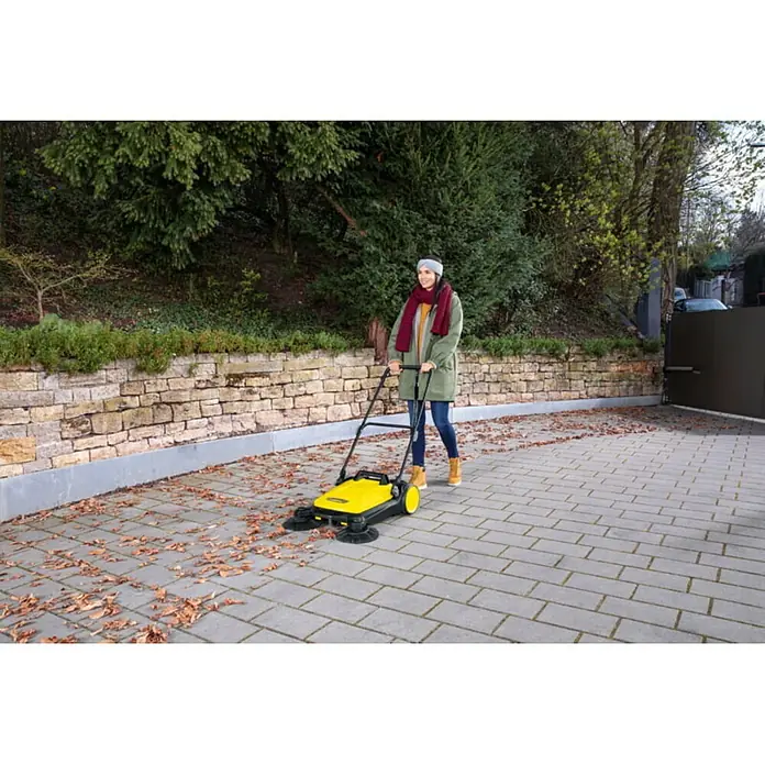Kärcher Barredora manual S 4 Twin - Volumen de recipiente: 20 l, Rendimiento por unidad de superficie: 2.400 m²/h - Una mujer está utilizando una barredora amarilla para quitar hojas de una entrada pavimentada.