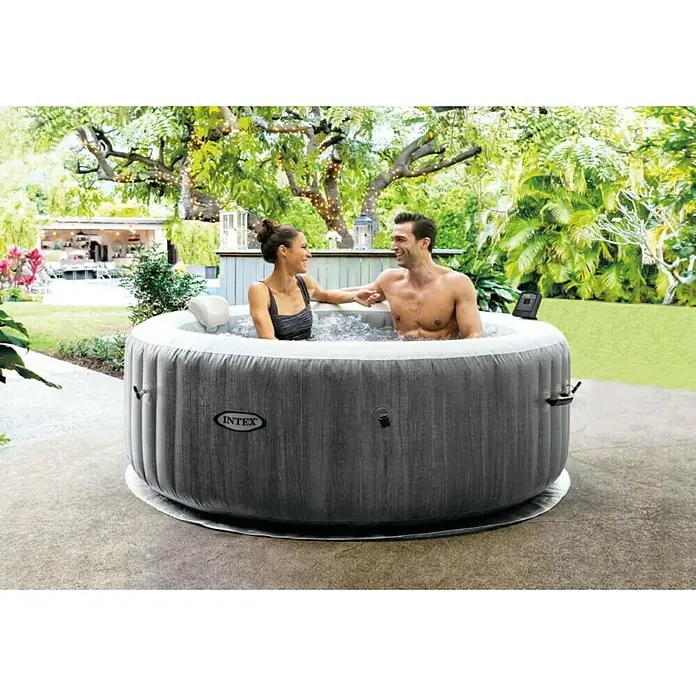 Intex Pure Spa Hidromasaje móvil Greywood Deluxe - Ø x Al: 196 x 71 cm, Gris/Blanco, 795 l - Bañera de hidromasaje Intex en el jardín con dos personas. La bañera de hidromasaje gris con aspecto de madera está en un patio e invita a relajarse.